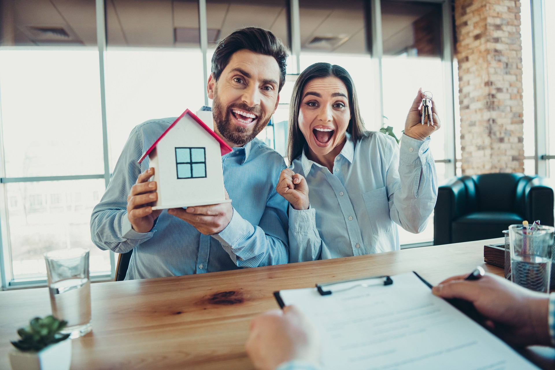 Excited young couple celebrates their new home purchase, showing house keys and a model, inside a bright workspace
